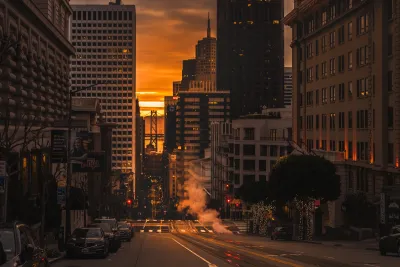 View down a San Francisco street with tall offive buildings on either side and a golden sunrise on the horizon.