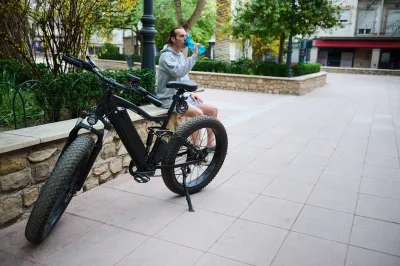 Black electric bike parked on urban walkway with rider sitting on ledge drinking water behind it.