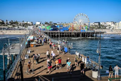 People walking on Santa Monica Pier boardwalk with Ferris wheel visible close to shore on sunny day.