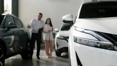 Close-up on white sedan in showroom with salesperson and client standing in background, blurred.