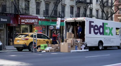 FedEx delivery truck with brown boxes stacked up behind it and yellow NYC van cab stopped at curb on NYC street.