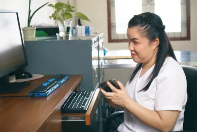 Visually impaired woman in white shirt sits at computer using screen reader device.