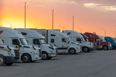 Row of mostly white and one red truck parked in a parking lot at sunset.