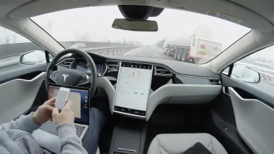 Interior view of man sitting in driver's seat of Tesla car holding laptop and mobile phone in lap.