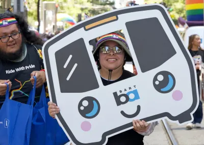 Person in bucket hat and sunglasses holding cardboard cutout of cartoon BART train in parade in San Francisco.