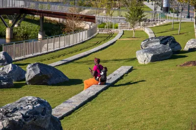 An African American woman with a baby on her back sitting in the park surrounded by lush green trees, grass and plants with blue sky at Rodney Cook Sr. Park in Historic Vine City in Atlanta, Georgia.