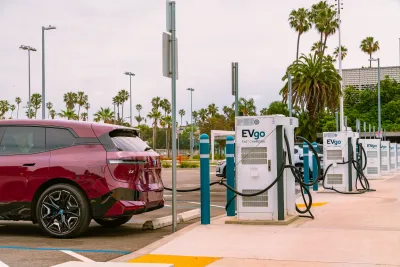 Red electric car plugged into charging station in outdoor parking lot with palm trees in background.