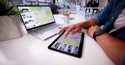 Person with laptop and tablet on desk looking at real estate listings.