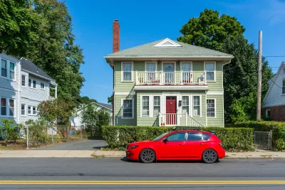 Green two-story wood house in Brighton, Massachusetts on a sunny day with small red sedan parked in front.