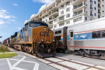 Amtrak train next to freight train at a station next to multi-story apartment building.