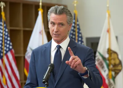 Gov. Gavin Newsom speaking behind a podium, pointing a finger upwards. The American and California flags are behind him.