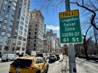 Green and yellow congestion peicing zone sign on post in NYC next to street with moderate traffic.