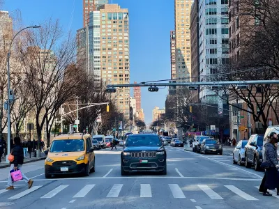 New York City crosswalk with a few pedestrians and yellow cab and black SUV stopped at light.