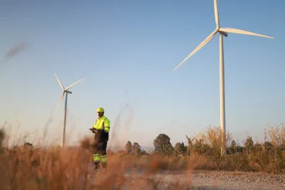 Worker in yellow safety jacket standing in field next to wind turbine.