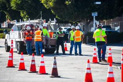 Group of workers in yellow and orange safety vests and hard hats stand in street in Los Angeles next to work truck protected by row of orange cones.