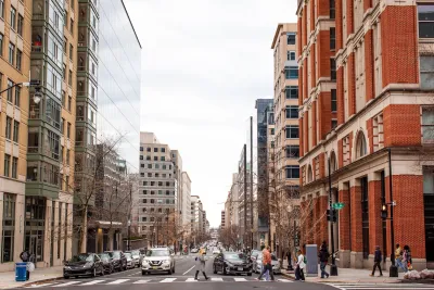 Street scene in Washington DC with mixed modern and brick buildings and pedestrians crossing a crosswalk.