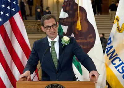 Daniel Lurie, in a suit and green tie, speaks behind a podium with the American flag in the background.
