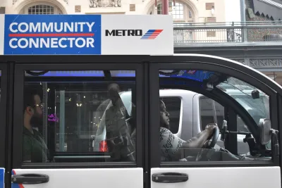 A modern Metro Community Connector (Cab) EV Vehicle, cruising during midday with a passenger in the downtown financial district area.