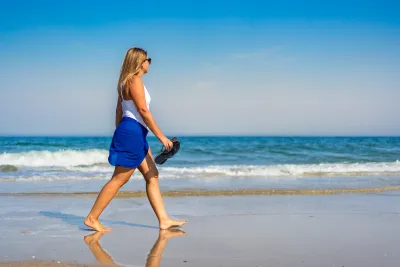 Woman in white top walks on beach holding her sandals on sunny day.