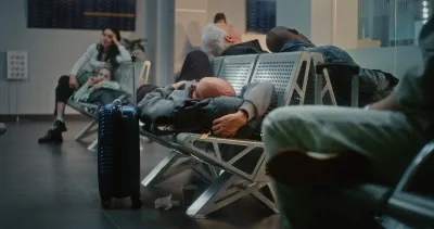 Man sleeping on airport seats in terminal with other people sitting around him.