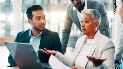 Man and woman in business meeting looking at laptop with another man leaning over desk.
