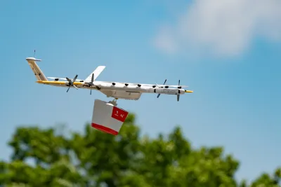 White drone holding red and white delivery box flying over green treetops.
