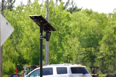 A black Flock traffic camera with solar panel above it installed on a pole outdoors with car visible in background.