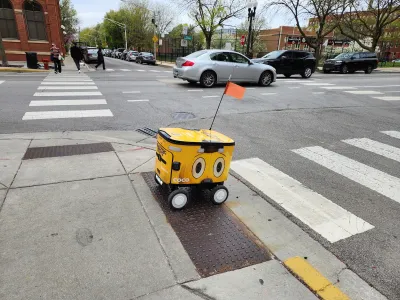 Yellow delivery robot rolling across curb cut onto painted crosswalk.