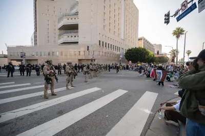 National Guard troops in uniform lined up on a street in downtown Los Angeles next to high-rise jail building with protesters facing them across the street.