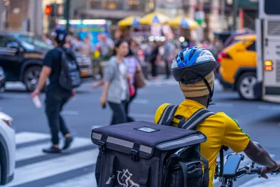 A person in NYC bikes behind a bus. They have a helmet on top of a yellow hat and wear a yellow shirt.