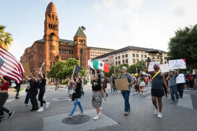 Protesters carrying signs and Mexican and American flags march in San Antonio street against ICE raids with historic Spanish-style building in background.