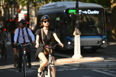 Woman in black wearing helmet on bicycle with man in white shirt and tie and helmet riding on bike behind her with bus visible in background in Paris, France.