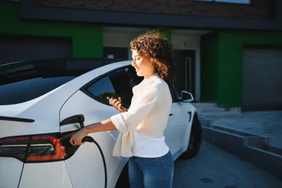 Woman in white shirt charging white electric sedan in front of house.