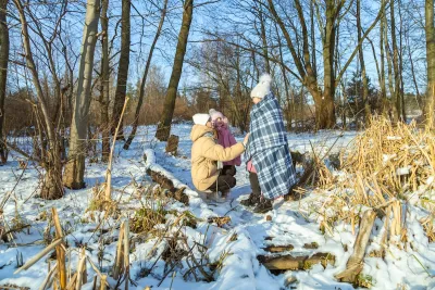 A woman in the snowy woods wraps two children in blankets.