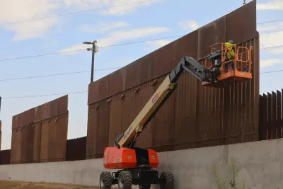 Border wall construction. A man in a yellow jacket is in a crane bucket.