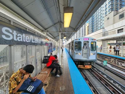Male passengers sit on wood bench at State and Lake Elevated Train Station with train approaching in Chicago.
