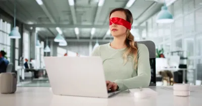 Woman wearing red blindfold sitting at white table typing on silver laptop.
