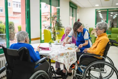 Three senior women sitting at table with caretaker leaning over them in bright group home.
