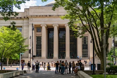 People walk in front of an MIT building with marble pillars. There are trees in the foreground.
