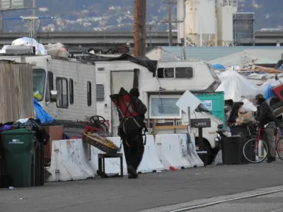 Man walks past homeless encampment behind concrete Jersey barriers on street in Oakland, California with RVs parked behind tents.
