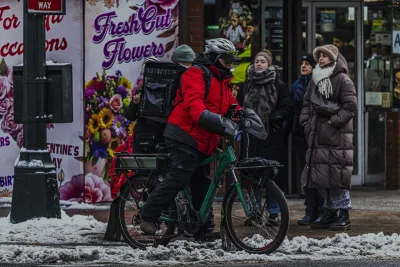 Man in red jacket on e-bike wearing black delivery backpack in winter on New York City street.