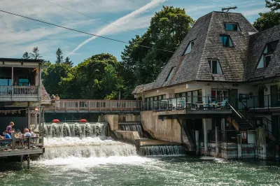 The Leeland Dam, nestled between two buildings with pointed roofs. People watch the water.