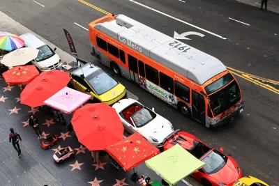 A Metro bus drives down a street on a rainy day. Parallel to the bus is a line of parked cars, then, a line of umbrellas. 
