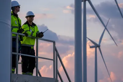Two construction workers on a wind turbine, with two more in the background against a pink sky.