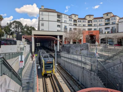 Yellow LA Metro train pulling out of station in Pasadena, California with neo-Spanish style apartment building visible above station.