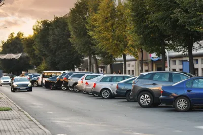 A row of cars parked next to a building. One car drives perpendicular to the parked cars.