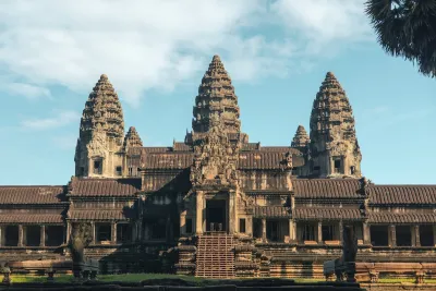 The Ancient Angkor Wat temple in Cambodia captured in early morning light. The temple is gray, and has three pillars with cone shaped tops. It is under a light blue sky.