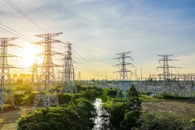 Many large power transmission towers loom over a small river with green trees on either side at sunset.