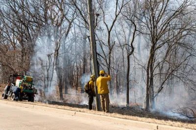 Two firefighters stand at edge of paved road looking at burned, smoldering forest after fire.