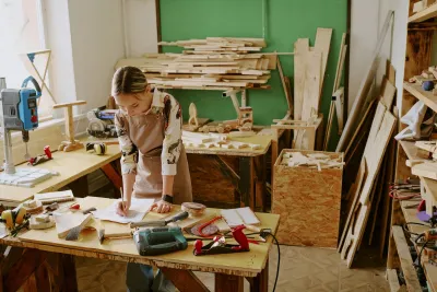 Woman leaning over work table in woodworking space surrounded by lumber.